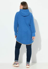 Blauer, flauschiger Hoodie mit einem abgerundeten Saum und orangefarbigen Akzenten. Er hat lange Ärmel und eine lockere Passform, kombiniert mit dunklen Jeans und bunten Turnschuhen.