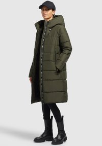 Mujer vestida con un abrigo largo acolchado en color verde oliva con capucha, leggings negros, botas negras y una gorra negra, de pie contra un fondo gris.