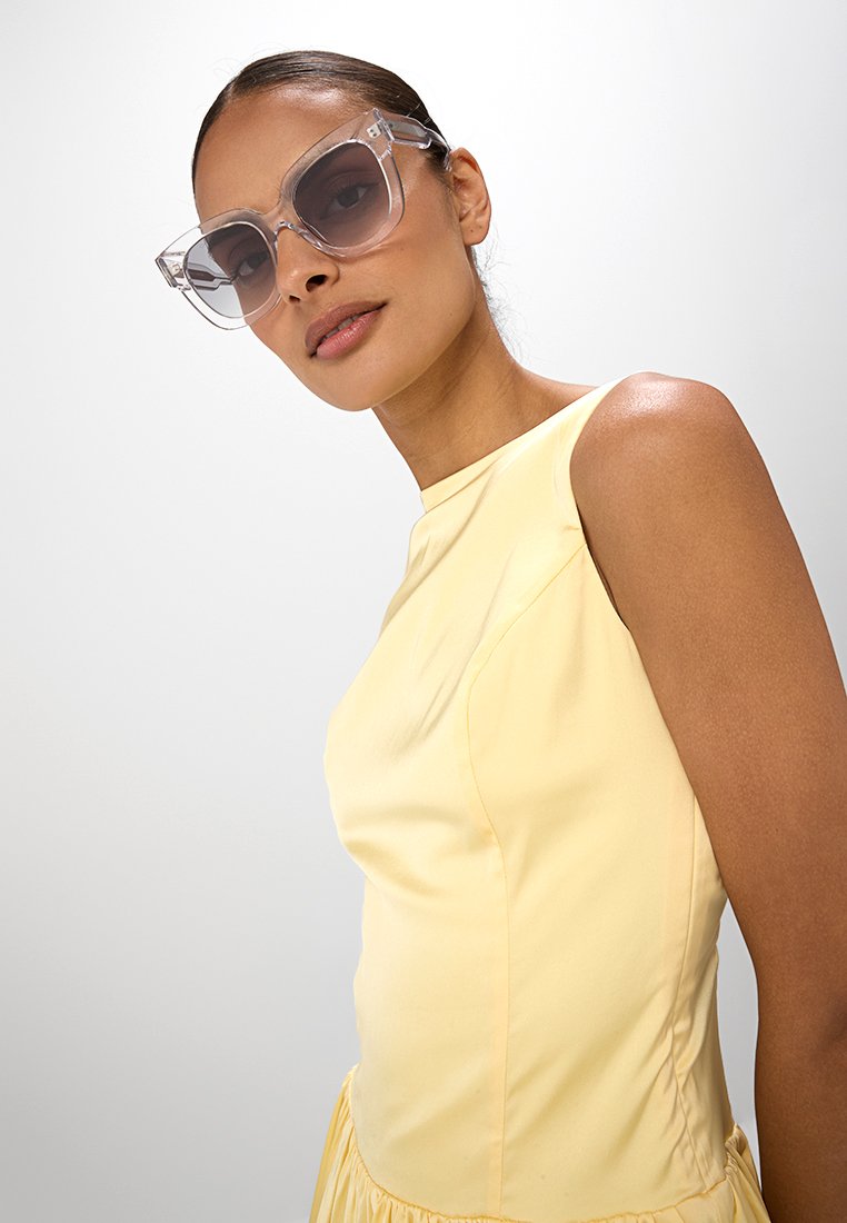 Gafas de sol grandes y transparentes con lentes marrones, con montura angular. El modelo lleva un vestido amarillo sin mangas con una textura suave.