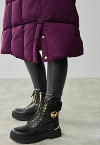 Personne portant des bottes noires en cuir à lacets avec des détails dorés et des leggings noirs sous un manteau matelassé violet avec des boutons-pression.