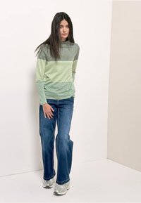Grün- und grau gestreiftes Langarmshirt, schlanke Passform, kombiniert mit ausgestellten blauen Jeans. Modell trägt silberne Sneakers und steht vor einem schlichten Hintergrund.