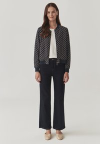 Chaqueta de bombardero navy con un patrón geométrico, puños y dobladillo acanalados. Combinada con pantalones de mezclilla oscuros de campana y zapatos de color claro.