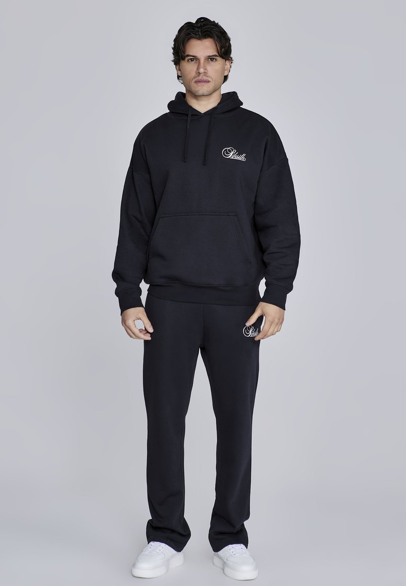 Črna kapuca z žepom spredaj in logotipom, združena s črnimi trenirkami. Iz bombaža, sproščen kroj in bele teniske za končni videz.