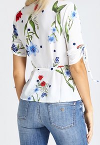 Weißes Blusenshirt mit floralen Mustern in Rot und Blau, mit kurzen Ärmeln und gebundenem Taillenzug, kombiniert mit blauen Jeans.
