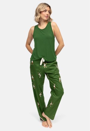 Vrouw met een groene mouwloze top en groene broek met tennisserprint, staand op blote voeten met handen achter de rug.