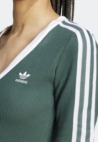 Zelený žebrovaný top s výstřihem do V, bílým lemováním a třemi vodorovnými bílými pruhy na rameni; obsahuje vyšitý bílý logo.