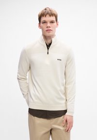 Pulover bej cu fermoar jumătate, realizat dintr-un material moale, având o textură ribbed pe umeri și un logo negru. Purtați pe un tricou maro închis.