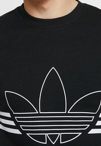 Svart sweatshirt med en framträdande vit Adidas trefoil-logotyp med tre blad ovanför horisontella ränder. Mjuk bomullstextur.