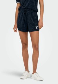 Marineblaue Velours-Shorts mit elastischem Bund und Kordelzug, mit "NYC" in weißem Text auf der linken Seite. Kombiniert mit weißen Turnschuhen.