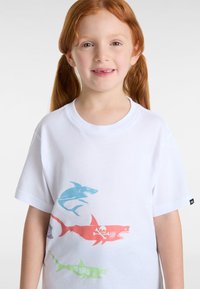 Weißes T-Shirt mit vier Haifischdesigns in Blau, Rot und Grün, sowie einem Grafikdesign von Totenkopf und gekreuzten Knochen. Aus weichem Baumwollstoff.