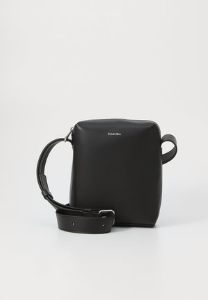 Sac en cuir noir en forme de carré, avec une bandoulière réglable et un logo en relief. Texture lisse et design minimaliste.
