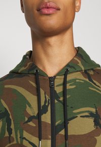 Sudadera con capucha en un patrón de camuflaje con tonos verdes, marrones y negros; cuenta con cierre de cremallera y detalles de cordón.