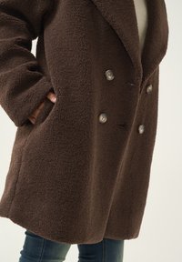 Pruun topeltööpekkega mantel, mis on valmistatud tekstureeritud fleece-kangast, millel on suured nööbid ja küljetaskud. Mantel on laia lõikega ja laiad kraed.