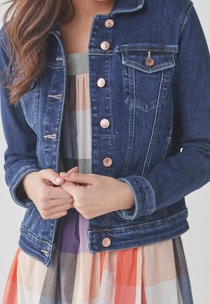 Femme boutonnant une veste en jean bleue par-dessus une robe à carreaux multicolores, montrée des épaules à la taille.