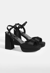 Schwarze, hochhackige Plateausandalen mit glänzender Oberfläche, offener Zehenpartie, dickem Blockabsatz, verstellbarem Riemen und Überkreuz-Design.