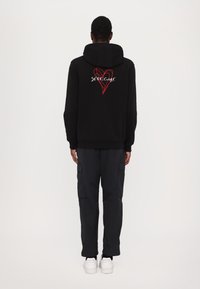 Haut à capuche noir avec un grand motif de cœur rouge et le texte "SETTE.CUORE" au dos. Associé à un pantalon cargo noir et des baskets blanches.