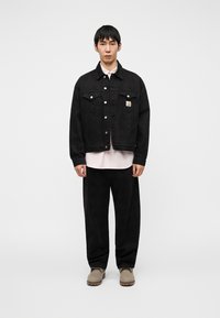 Μαύρο τζιν μπουφάν με μπροστινές τσέπες και κουμπιά τύπου κλικ, φορεμένο πάνω από ένα ανοιχτό ροζ πουκάμισο και μαύρο φαρδύ παντελόνι, σε συνδυασμό με μπεζ μπότες.