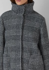 Szary płaszczyk w kratę z wysokim kołnierzem, miękką teksturą i dwoma bocznymi kieszeniami, z mieszanką czarnych i białych wzorów na całej powierzchni.