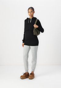 Pregnant woman stands wearing a black hoodie, gray sweatpants, brown platform shoes, and carries an olive green shoulder bag.