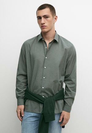Giovane uomo con capelli corti che indossa una camicia verde a quadri con bottoni e un maglione verde scuro legato intorno alla vita.
