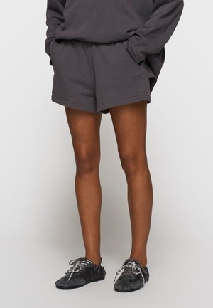 Persoon draagt donkergrijze casual shorts en bijpassende sweatshirt met handen in de zakken, gecombineerd met zwarte en grijze vetersneakers.