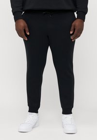 Schwarze Jogginghose in schmaler Passform, aus weichem Stoff mit elasticem Bund und gerippten Knöcheln, kombiniert mit weißen Turnschuhen.