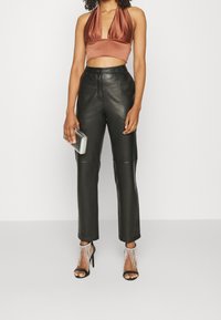 Top corto in satin marrone a halter, pantaloni neri in pelle a vita alta e tacchi neri con dettagli in catena d'argento. Clutch argentata in mano.
