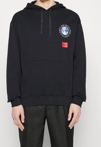 Sweatshirt à capuche noir en coton. Présente une poche avant, un cordon de serrage pour la capuche, et un imprimé graphique avec du texte sur la poitrine. Étiquette logo rouge sur la poche.