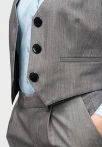 Γκρι υφασμάτινο γιλέκο κοστουμιού με τέσσερα μαύρα κουμπιά και μια τσέπη, σε συνδυασμό με ανοιχτό μπλε πουκάμισο από κάτω. Ομαλή υφή, ραμμένο σε εφαρμοστή γραμμή.