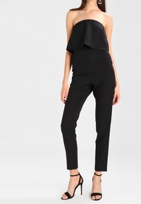 Czarny kombinezon bez ramiączek z dopasowanym tułowiem i luźnym, rozszerzanym overlayem. Posiada zwężające się nogawki i wysokie szpilki. Gładka, jedwabiście gładka tekstura.