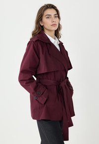 Bordowy płaszcz trencz z szerokim kołnierzem, paskiem w talii i zapinanymi na guziki mankietami. Gładka tkanina o fasonie dopasowanym, z warstwową konstrukcją z przodu.