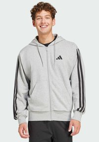 Šedá mikina na zips z mäkkého materiálu. Má čierne pruhy na rukávoch, predné vrecko a kapucňu so šnúrkami. Na hrudi sa nachádza logo Adidas.