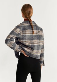 Rutet jakke i marineblå, cremefarvet og brune nuancer; har en krave, sænket skuldre og knaplukning ved ærmerne; parret med sorte leggings.