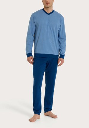 Uomo che indossa una maglia a maniche lunghe blu con scollo a V e pantaloni abbinati blu navy, in piedi a piedi nudi su uno sfondo chiaro.