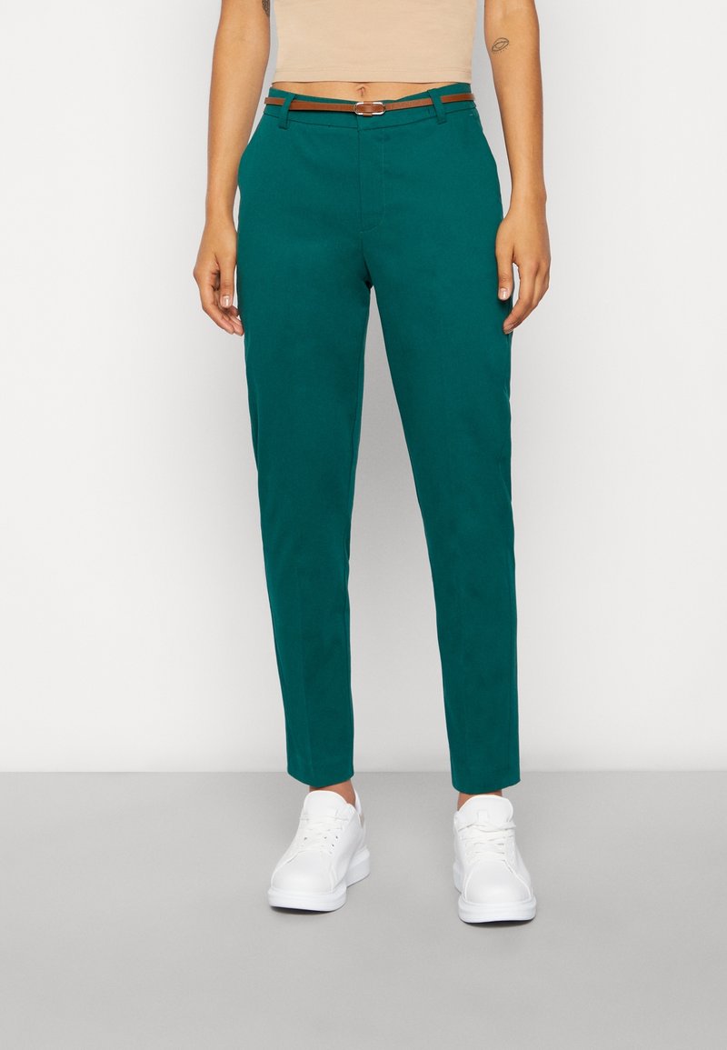 Tyrkysové kalhoty s úzkým střihem a hladkým povrchem, hnědý pásek akcentující pas, kombinované s bílými teniskami, zobrazené na neutrálním pozadí.