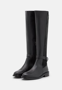 Bottes noires montantes en cuir lisse et en daim, avec une fermeture éclair sur le côté et une sangle ajustable avec une boucle en métal.