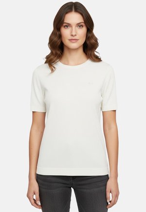 Donna con capelli castani ondulati indossa una maglietta bianca a maniche corte e jeans scuri, in piedi davanti a uno sfondo bianco, rivolta verso la fotocamera.