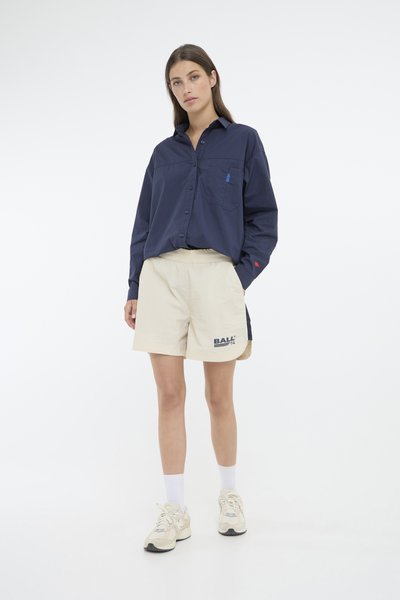 Marineblå skjorte med knapp foran og brystlomme, beige shorts med grafisk trykk, og lyse sneakers. Modellen står foran en nøytral bakgrunn.