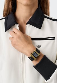 Skaitmeninis rankinis laikrodis su auksiniu stačiakampiu ciferblatu, juodu silikono dirželiu ir skaitmeniniu ekranu, rodančiu laiką. Nešiojamas ant rankos.