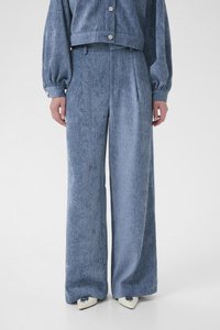 Denim wide-leg pants in light blue, featuring a textured finish, front pleats, and a relaxed fit, paired with pointed white shoes.