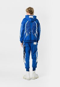 La sudadera azul con capucha y los pantalones de chándal a juego presentan diseños de alas en blanco y azul. La sudadera con capucha tiene grandes gráficos de alas en la parte posterior y en las mangas.