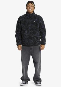 Schwarzer Fleece-Pullover mit lila wirbelndem Muster, 1/4 Reißverschluss, kombiniert mit baggy grauen Jeans und schwarz-weißen Sneakers.