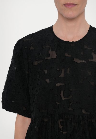Black blouse featuring textured floral patterns, short sleeves, and a round neckline. Sheer overlay with layered ruffles at the hem.