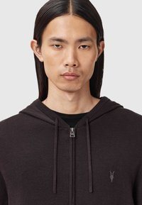 Ung man med rakt långt hår som bär en mörk zip-up hoodie med ett litet broderat emblem på bröstet, neutral bakgrund.