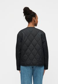 Sort quiltet jakke med diamantmønster, rund halsudskæring og lange ærmer, stylet med lyseblå jeans. Ingen synlige lukninger.