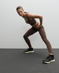 Brun sportbehå och matchande högmidjade leggings, tillverkade av slät, elastisk tyg. Svarta träningsskor med ett texturerat mönster.