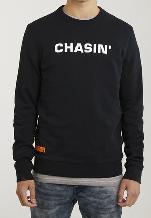 Mann trägt schwarzen Pullover mit weißem "CHASIN'"-Schriftzug, grauem Hemd darunter und ausgewaschenen blauen Jeans, sichtbar vom Hals bis zur Hüfte.