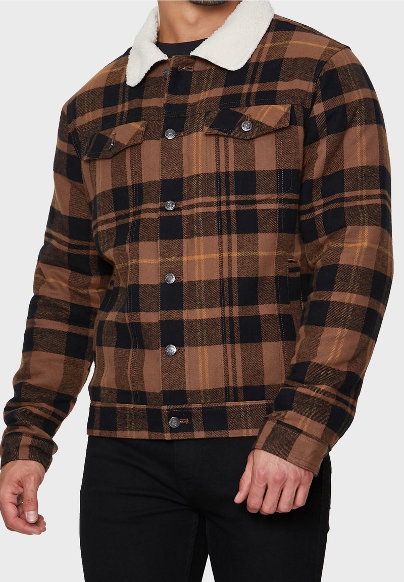 Veste à carreaux marron et noir avec un col en peau lainée couleur crème, fermeture à boutons et deux poches poitrine. Tissu texturé avec des accents jaunes.