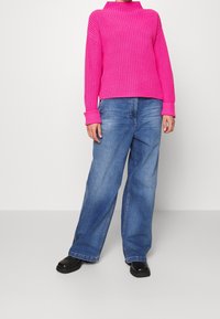 Maglione a coste fucsia con colletto alto, abbinato a pantaloni di jeans denim a gamba larga di un blu chiaro. Calzature nere con suola spessa completano il look.