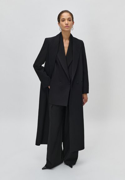 Donna con capelli intrecciati indossa un lungo cappotto nero, blazer doppiopetto, pantaloni a gamba larga e scarpe nere appuntite, in piedi davanti a uno sfondo grigio.